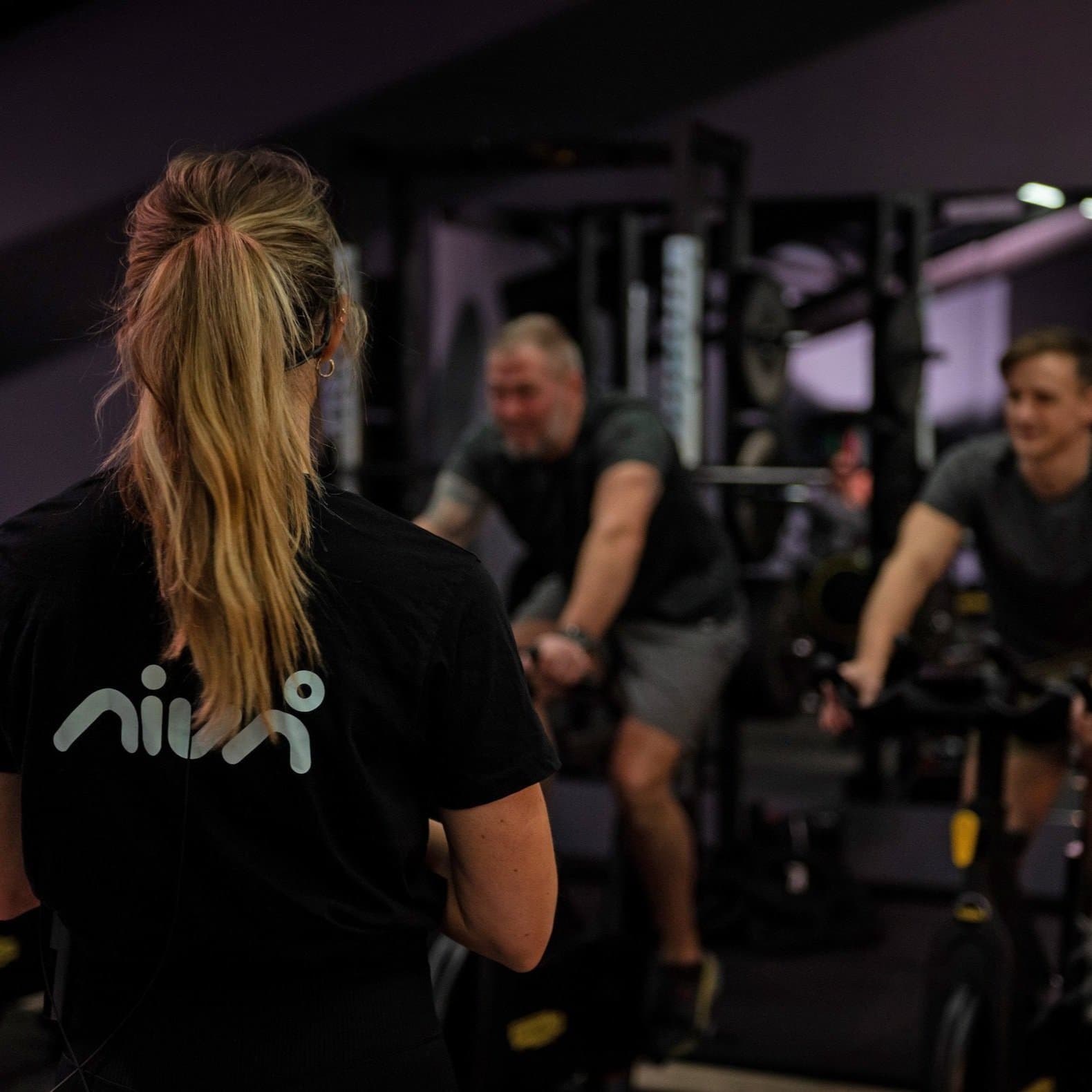 An instructor in a Nivå shirt in front of two members on spin bikes at Nivå Trening in Halden.
